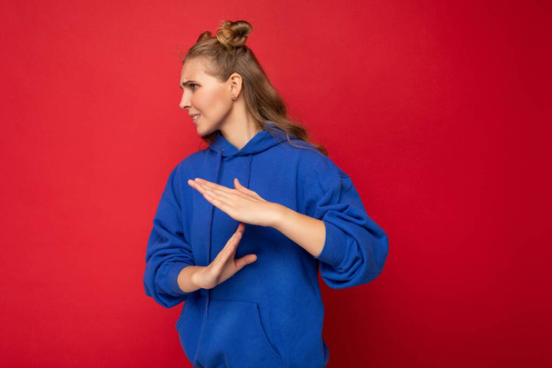 Photo of emotional young attractive blonde woman with two horns with sincere emotions wearing hipster bright blue hoodie isolated over red background with copy space and showing pause sign - Photo, Image