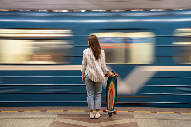 Chica con longboard de pie en la plataforma subterránea con borroso tren azul en movimiento en el fondo - Foto, imagen