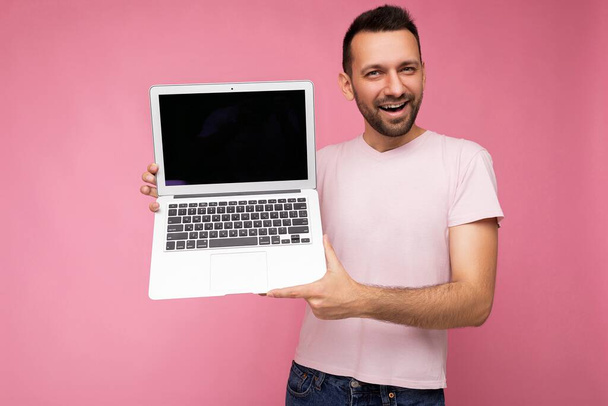 Guapo sonriente morena hombre sosteniendo ordenador portátil mirando a la cámara en camiseta sobre fondo rosa aislado - Foto, imagen