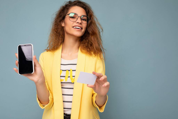 Portret van een gelukkige mooie vrouw met een geel jasje en een optische bril geïsoleerd op de achtergrond muur vasthouden en tonen telefoon met leeg scherm en creditcard op zoek naar camera - Foto, afbeelding