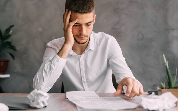Migraine and headache in a young man in a bank or an office worker sitting at a workplace with documents. The guy looks at the bankruptcy documents. Job search and resume. Work or business concept - Photo, Image