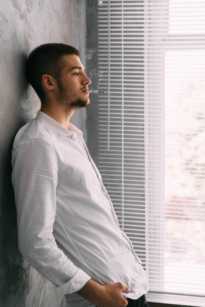 office police worker standing by the window smokes a cigarette blowing out smoke. Serious Caucasian man smokes in the workplace. Addiction is bad habit. Tobacco smoking. Health and poisoning concept - Photo, Image