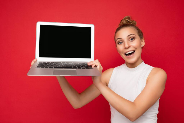 Happy amazed smiling beautiful blond young woman with gathered hair looking at camera holding computer laptop with empty monitor screen with mock up and copy space wearing white t-shirt isolated on - Fotó, kép