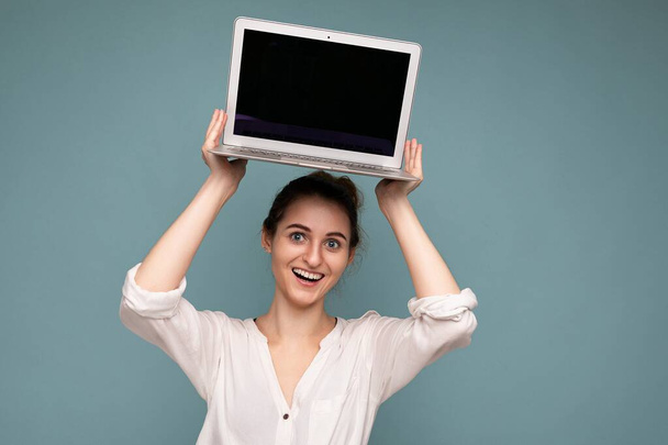 Kaunis hymyilevä nuori nainen jolla netbook tietokone katselee kameraa yllään valkoinen paita eristetty sinisellä pohjalla - Valokuva, kuva
