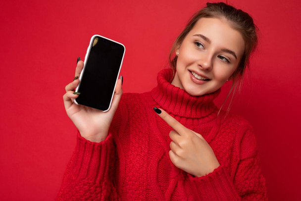 Фото красивой милой улыбчивой молодой женщины в теплом красном свитере, изолированном на красном фоне стены, держа смартфон и показывая телефон с пустым дисплеем для макета глядя в сторону и - Фото, изображение