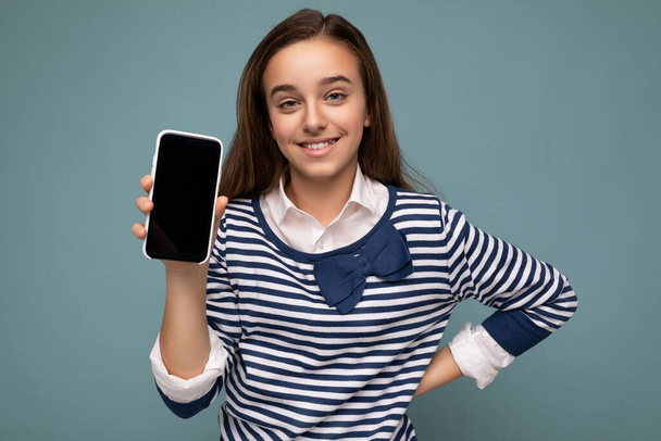 Pretty positive smiling girl wearing striped longsleeve standing isolated on blue background with copy space holding smartphone showing phone in hand with empty screen display for mockup pointing at - Foto, Bild