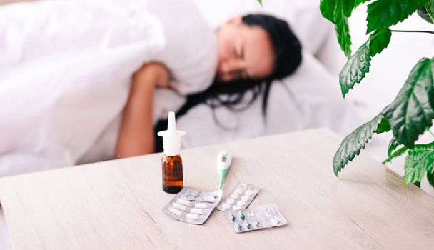 una mujer joven que sufre de gripe o fiebre estacional, el concepto de tratamiento, pastillas y medicamentos en la mesita de noche con un gran plano - Foto, imagen