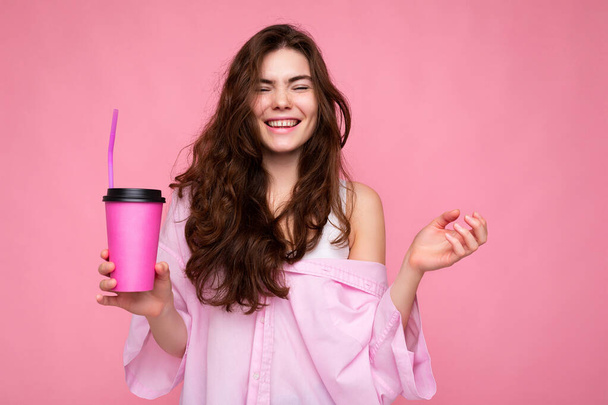 Beautiful young positive brunette woman wearing casual stylish clothes isolated over colorful background wall holding paper cup for mockup drinking coffee looking at camera - Fotografie, Obrázek