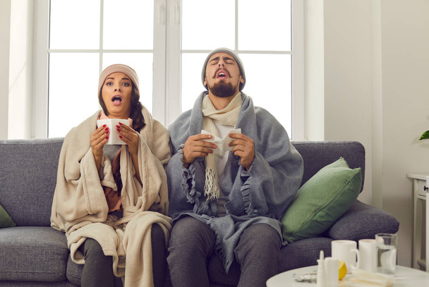 Sick young couple wrapped in blankets sitting on sofa and sneezing in paper tissues - Photo, image