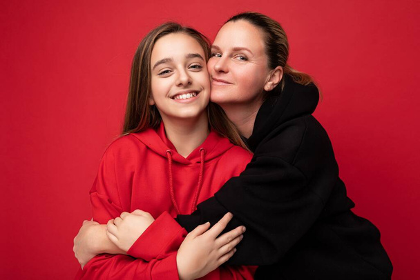 Shot of beautiful happy smiling little daughter wearing stylish red hoodie and adult mother wearing trendy black hoodie standing isolated over red background wall looking at camera and having fun - Photo, Image