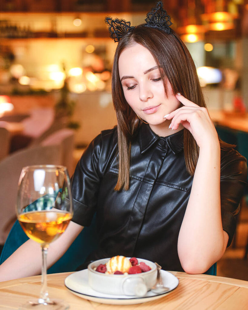 Portrait of young gorgeous woman drinking champagne in glass and looking with smile eating ice cream fruit dessert. Enjoying her leisure time inside a restaurant. Girl in a black dress with cat ears. - 写真・画像