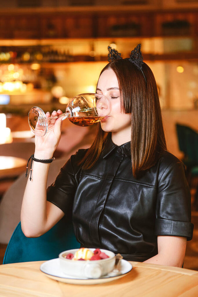 Portrait of young gorgeous woman drinking champagne in glass and looking with smile eating ice cream fruit dessert. Enjoying her leisure time inside a restaurant. Girl in a black dress with cat ears. - Foto, imagen