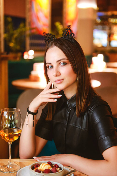 Portrait of young gorgeous woman drinking champagne in glass and looking with smile eating ice cream fruit dessert. Enjoying her leisure time inside a restaurant. Girl in a black dress with cat ears. - Fotografie, Obrázek