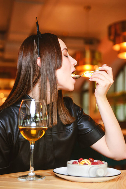 Portrait of young gorgeous woman drinking champagne in glass and looking with smile eating ice cream fruit dessert. Enjoying her leisure time inside a restaurant. Girl in a black dress with cat ears. - Photo, image