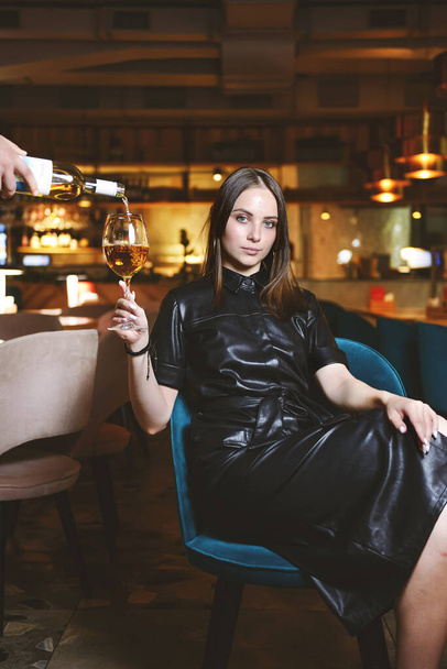 Portrait of young gorgeous woman drinking champagne in glass and looking with smile, enjoying her leisure time inside a restaurant. Girl in a black dress with cat ears. - Fotoğraf, Görsel