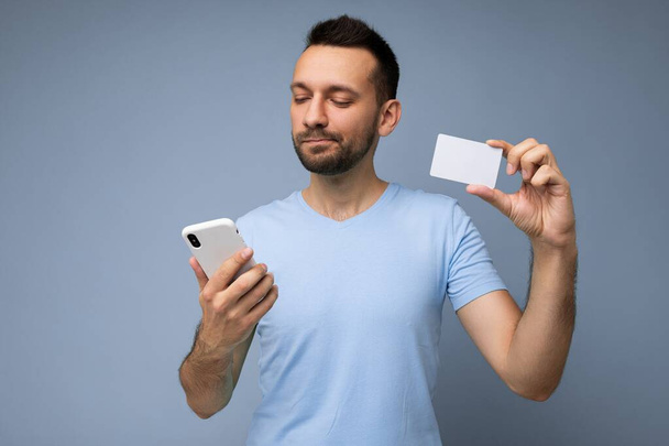 Arkadaki duvarda izole edilmiş günlük giysiler giyen yakışıklı bir adam telefon ve kredi kartı kullanıyor ve akıllı telefon ekranına bakarak ödeme yapıyor. - Fotoğraf, Görsel