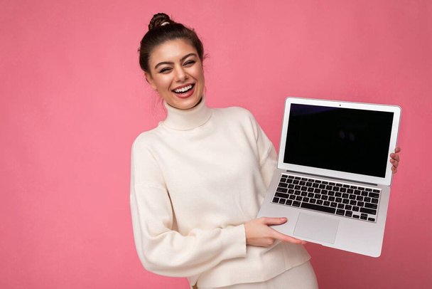 Photo of beautiful smiling and laughung brunette young lady with gathered dark hair wearing white sweater holding computer laptop and looking at camera with close eyes and open mouth isolated on pink - Photo, Image