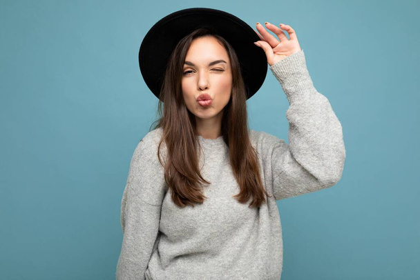 Young beautiful brunet woman. Trendy lady in casual grey sweater and stylish black hat. Positive female shows facial sincere emotions. Funny model isolated on blue background with free space giving - Φωτογραφία, εικόνα