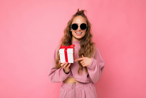 Atraktivní šťastný úsměv mladé blondýny kudrnaté ženy izolované přes růžové pozadí zdi na sobě růžové sportovní oblečení a sluneční brýle drží dárkové krabice při pohledu na kameru - Fotografie, Obrázek