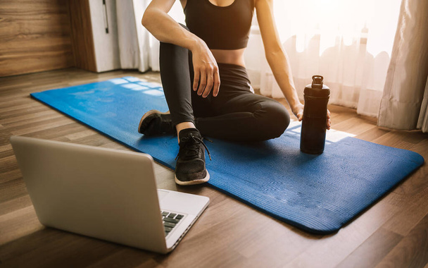 A sporty woman in sportswear is sitting on the floor with dumbbells and a protein shake or a bottle of water and is using a laptop. Sport and recreation concept - Фото, изображение