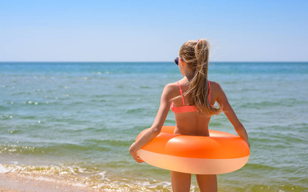 Dziewczyna z długimi włosami stoi na plaży z pomarańczowym gumowym pierścionkiem i patrzy na morze - Zdjęcie, obraz