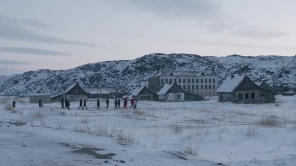 Turistas vai ao longo da rua do assentamento Teriberka passado casas abandonadas e destruídas contra o pano de fundo de rochas nevadas - Filmagem, Vídeo