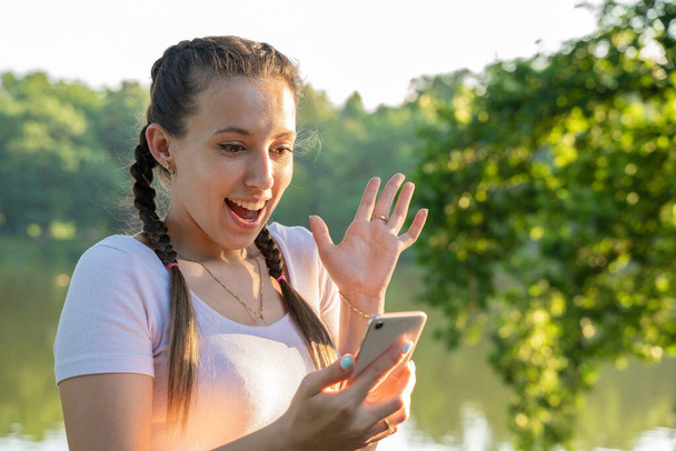 Девушка телефон. Эйфория кричит молодая девушка со смартфоном, показывая победный жест в солнечный день в парке. Мессинг, с удовольствием читающий текст - Фото, изображение