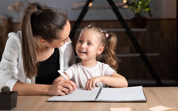 Caucasian young mother and little daughter sitting at the table in the evening, study online together, mom and little girl write by hand, do homework, study at home, online home schooling concept. - Foto, afbeelding