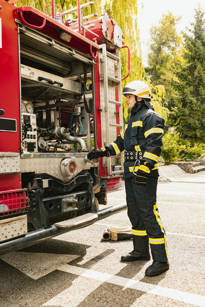 Один молодий чоловічий пожежник одягнений в уніформу з захисним шоломом біля пожежного двигуна. Вид збоку
 - Фото, зображення