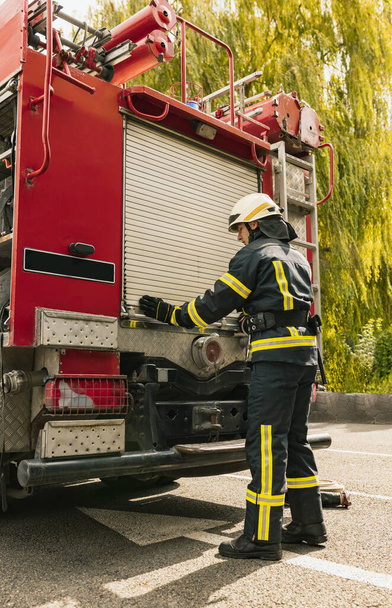 Один молодий чоловік пожежник одягнений в уніформу з захисним шоломом біля пожежного двигуна
. - Фото, зображення