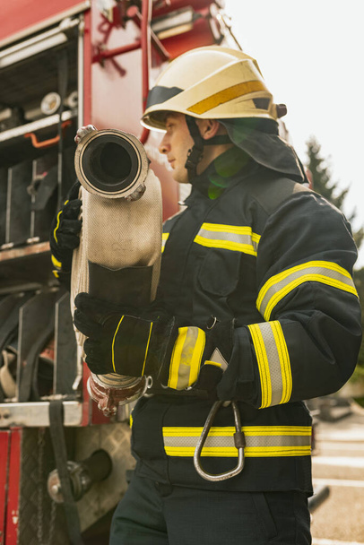 Один молодий чоловік пожежник, одягнений в уніформу з водяним шлангом перед пожежною машиною. Крупним планом
 - Фото, зображення