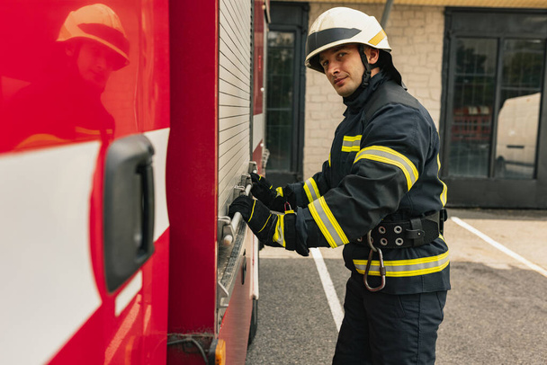 Ένας νεαρός άνδρας πυροσβέστης ντυμένος με στολή με προστατευτικό κράνος κοντά σε πυροσβεστική μηχανή. - Φωτογραφία, εικόνα