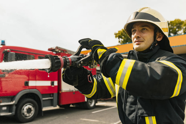 Ein junger Feuerwehrmann in Uniform mit Wasserschlauch löscht Brand - Foto, Bild