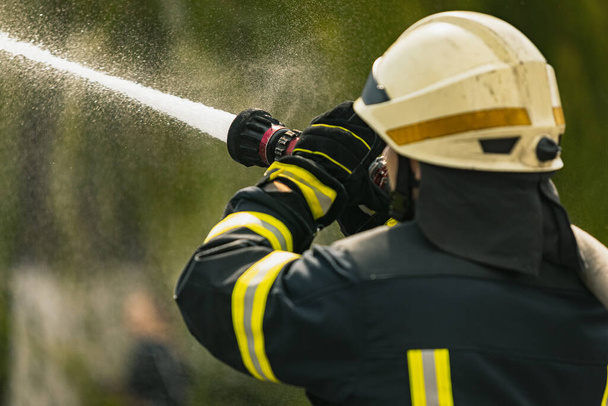 Un joven bombero de servicio vestido de uniforme con manguera de agua extinguiendo el fuego - Foto, imagen