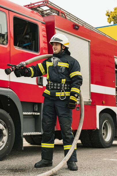 One young male firefighter dressed in uniform with water hose at fire truck ready for deployment - Foto, afbeelding