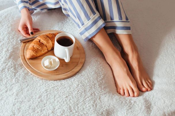 γυναίκες όμορφα πόδια σε πιτζάμες και δίπλα σε έναν καφέ με ένα κρουασάντ.την έννοια του καφέ στο κρεβάτι  - Φωτογραφία, εικόνα