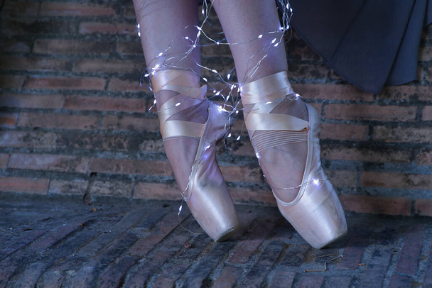 Szczegóły nóg i stóp kobiety klasycznej tancerki baletowej oświetlone paskami małych diodowych świateł, noszących brązowy kostium gazy na ceglanej ławce. - Zdjęcie, obraz