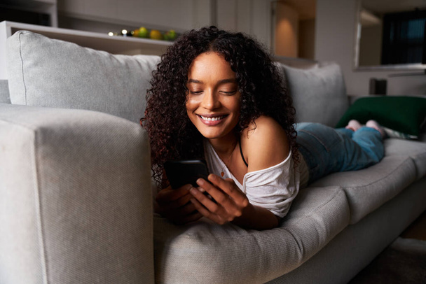 Mixed race female student relaxing on couch texting on cellular device - Fotó, kép
