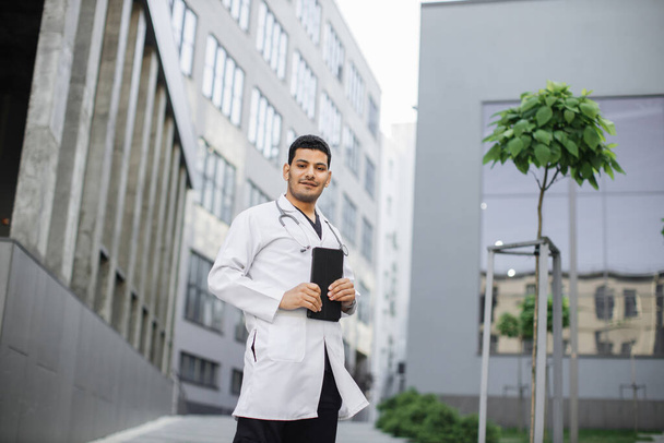 Male handsome Indian Asian doctor, working with patients records on digital tablet standing outside - Valokuva, kuva