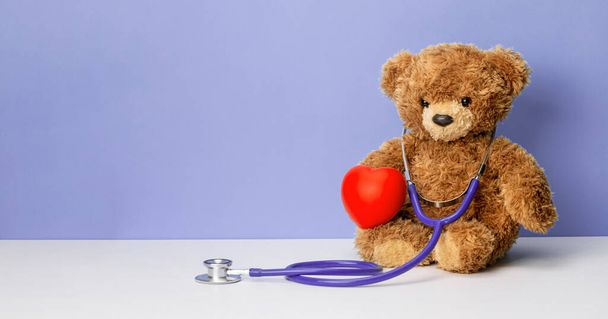 テディは紫色の背景に聴診器と心臓を持っている。家族の医者か小児科医の概念。テンプレートテキストのコピースペース - 写真・画像