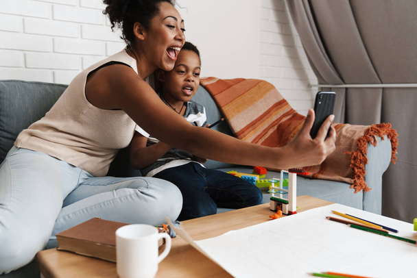 Black smiling woman taking selfie photo on cellphone while hugging her son at home - Foto, imagen