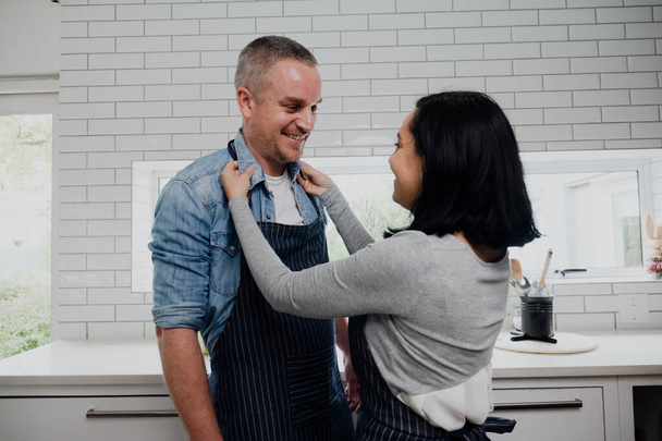 Usmívající se pár vaření v kuchyni, zatímco manželka dává zástěru na manžela. - Fotografie, Obrázek