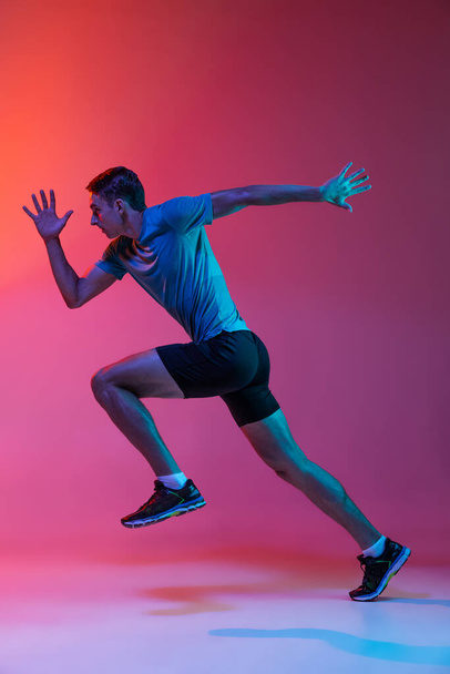 Portrat of Caucasian professional male athlete, runner training isolated on pink studio background with blue neon filter, light. Muscular, sportive man. - Valokuva, kuva