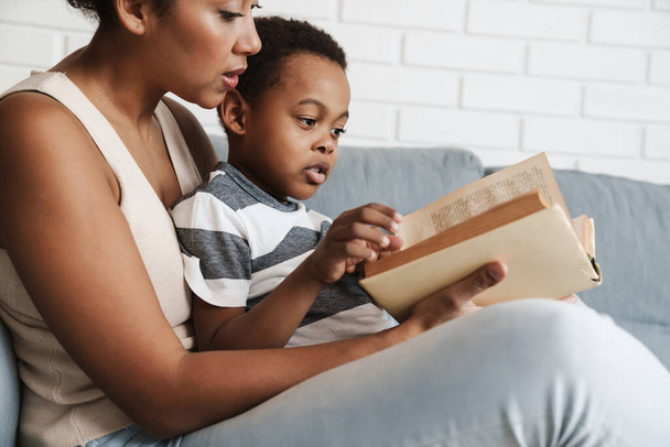 Black mother and son reading book while sitting on sofa at home - Foto, Imagem
