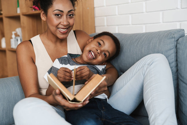 Black smiling mother and son reading book while sitting on sofa at home - Фото, изображение