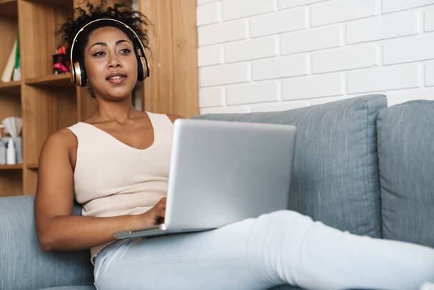 Femme noire dans les écouteurs travaillant avec un ordinateur portable tout en étant assis sur le canapé à la maison - Photo, image