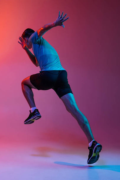 Portrat of Caucasian professional male athlete, runner training isolated on pink studio background with blue neon filter, light. Muscular, sportive man. Back view - Valokuva, kuva