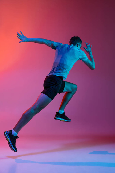 Retrato de atleta masculino profesional caucásico, entrenamiento de corredor aislado sobre fondo de estudio rosa con filtro de neón azul, luz. Hombre musculoso y deportivo. - Foto, imagen