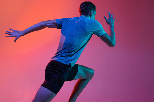 Portrat di atleta maschio professionista caucasico, allenamento corridore isolato su fondo rosa studio con filtro al neon blu, luce. Muscoloso, sportivo. Vista posteriore - Foto, immagini