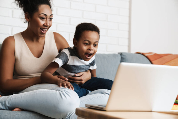 Black excited mother and son using laptop while sitting on sofa at home - Foto, Imagem